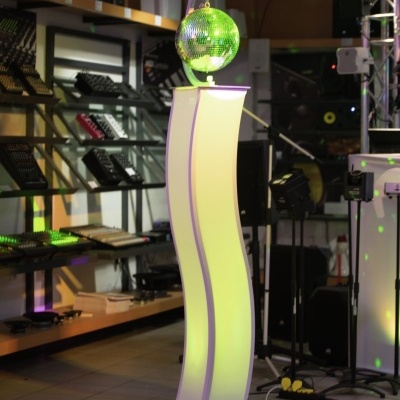 Coluna branca curva iluminada com esfera disco prateada refletindo luz verde em interior com equipamento de som.