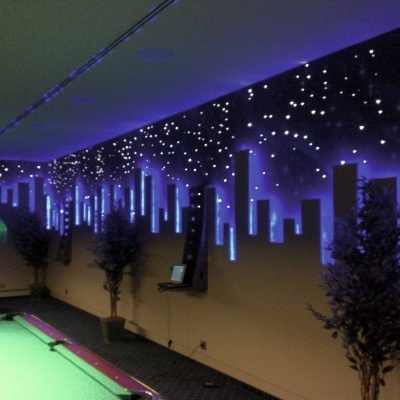 Mesa de bilhar verde numa sala com iluminação azul e decoração de parede temática de cidade e estrelas.