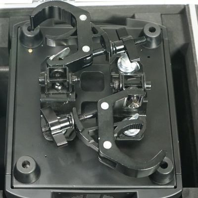 Dispositivo mecânico preto com garras ajustáveis numa base quadrada preta dentro de uma caixa cinzenta