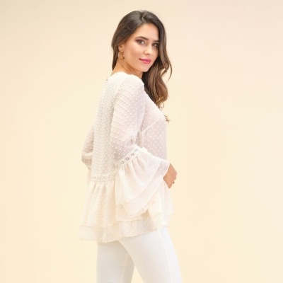 Mulher com blusa branca rendada de mangas largas e calças brancas em fundo bege.
