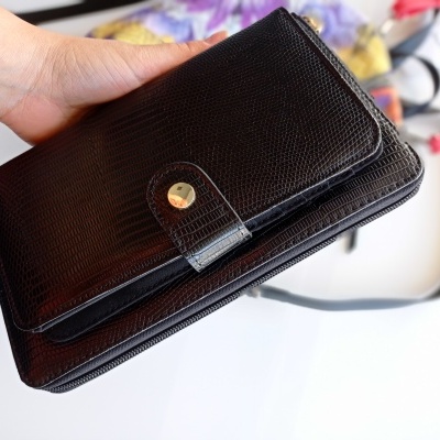 Carteira preta em pele com textura e fecho dourado, segurada à mão sobre fundo branco