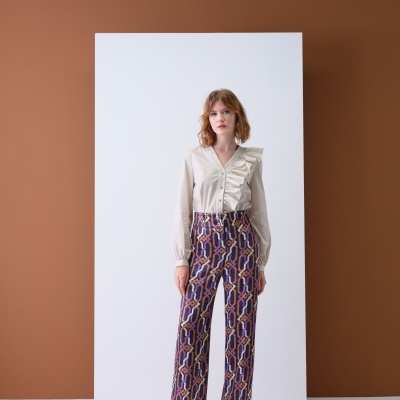 Modelo a vestir calças com padrão geométrico colorido e camisa bege com folho, calçado castanho, estúdio com fundo branco e castanho.