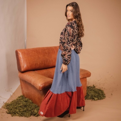 Mulher com saia comprida azul e vermelha e blusa floral preta em estúdio com poltrona castanha