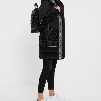 Mulher com casaco preto comprido com padrões florais, legging preta, sapatilhas brancas e mochila preta