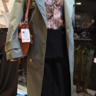 Manequim com casaco verde, camisa estampada castanha e preta, calças pretas e mala castanha com etiqueta