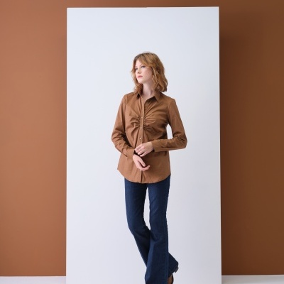 Mulher vestindo camisa castanha e calças azuis, em fundo branco e parede castanha.