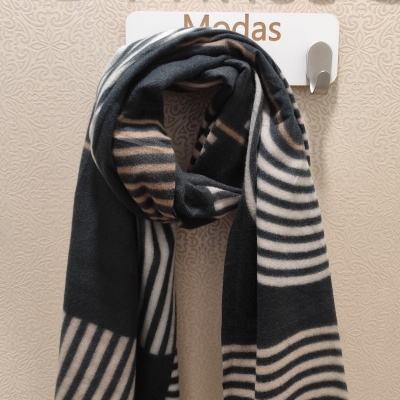 Cachecol listrado preto, bege e branco pendurado num gancho contra parede texturizada bege com placa e letras decorativas douradas.