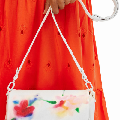 Mala branca com padrão floral colorido e alça branca, sendo segurada por mão sobre vestido laranja bordado