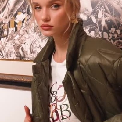 Mulher vestindo casaco verde acolchoado sobre t-shirt branca com letras, sentada em sofá com fundo decorativo