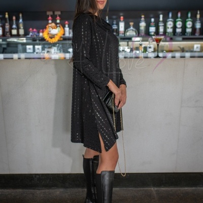 Mulher vestindo vestido preto com lantejoulas e botas pretas de salto alto, segurando mala pequena numa mão, com bar ao fundo.