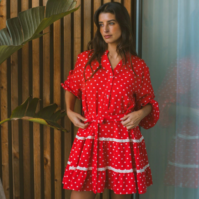 Vestido curto vermelho de bolinhas brancas com cinto e detalhes em renda numa mulher junto a parede de madeira e planta.