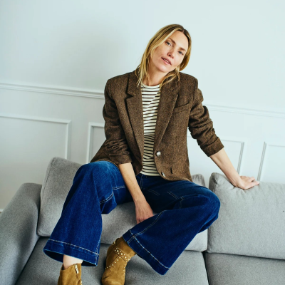 Mulher sentada num sofá a usar casaco castanho, camisola às riscas, jeans azuis largos e botas castanhas com tachas.