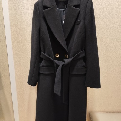 Casaco comprido preto com cinto e botões dourados pendurado em cabide.