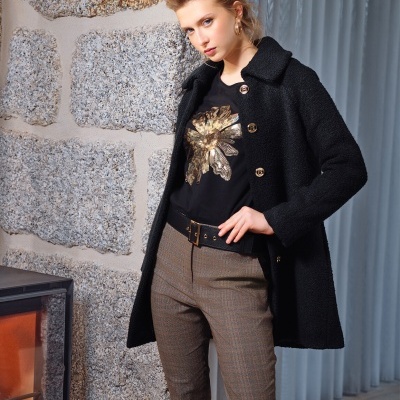 Mulher com casaco preto, t-shirt preta com estampa dourada, calças castanhas xadrez, posando perto de parede de pedra e lareira.