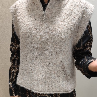 Colete de malha cinza claro com fecho de correr sobre camisa preta estampada e calças bege.