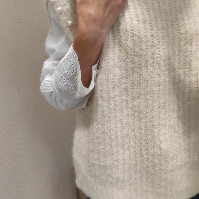 Colete de malha bege com pérolas no ombro, camisa branca bordada e calças jeans escuras