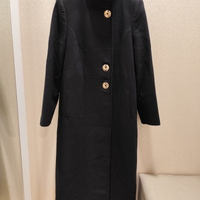 Casaco preto comprido com botões castanhos pendurado em cabide contra parede bege