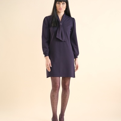 mulher vestindo vestido roxo escuro com laço, collants pretos de bolinhas e sapatos de salto preto em fundo bege