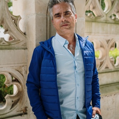 Homem com casaco azul acolchoado e camisa azul clara encostado em muro de pedra ornate