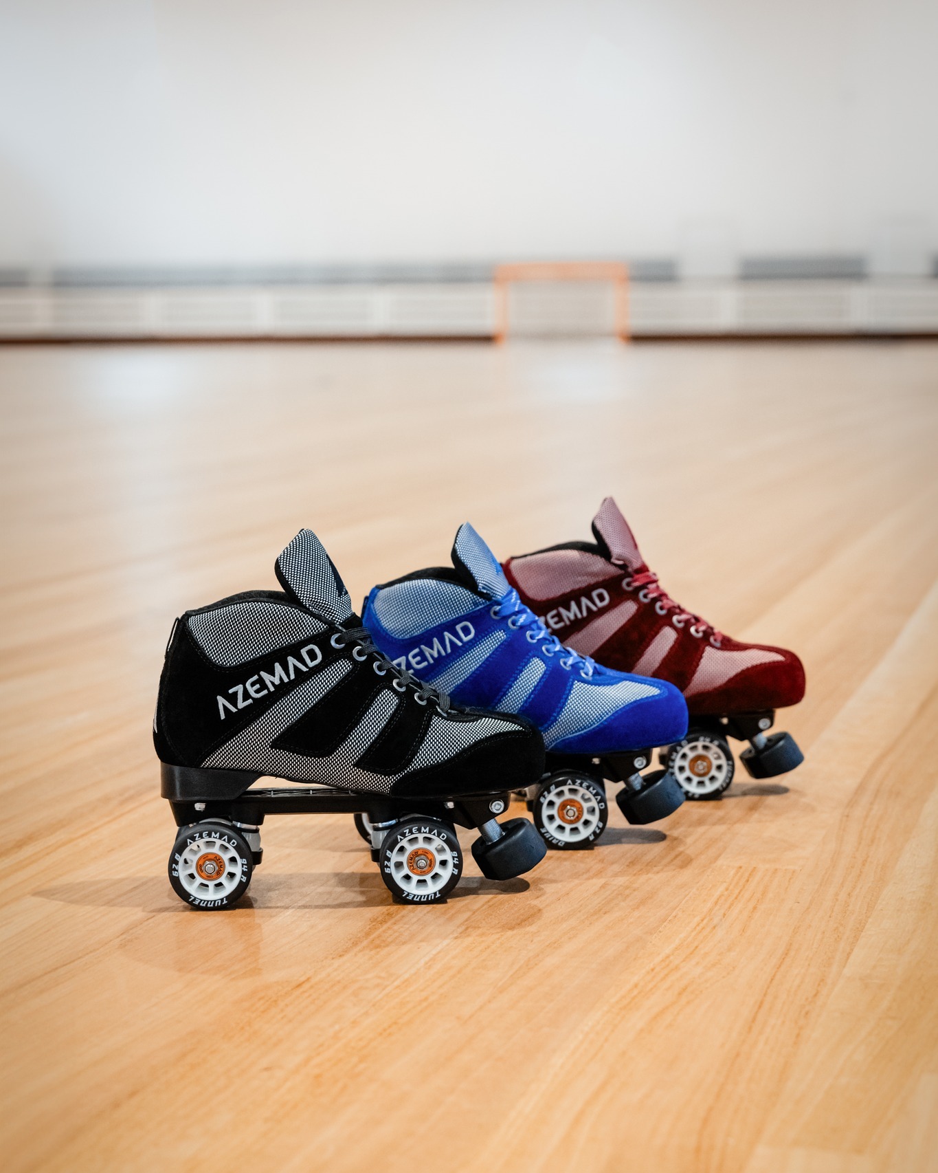 Três patins de quatro rodas nas cores preta, azul e vermelha, com texto AZEMAD, em chão de madeira