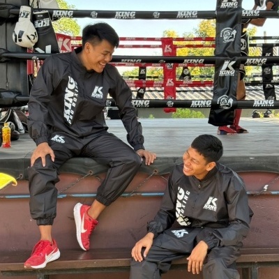 Dois homens com fato de treino preto KING PRO BOXING sentados e rindo numa bancada em ringue de boxe ao ar livre
