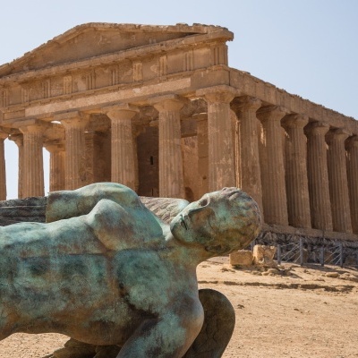 Escultura de homem deitado em bronze com templo grego antigo ao fundo