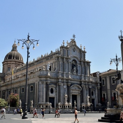 Praça com edifício histórico grande, fonte com escultura de leão e pessoas a caminhar