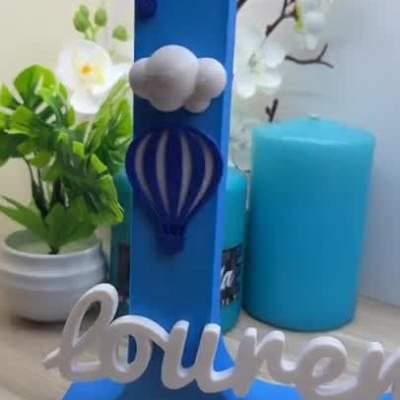 Peça decorativa com letra R azul, balões e nuvens, nome Rodrigo Lourenço em letras brancas sobre mesa de madeira