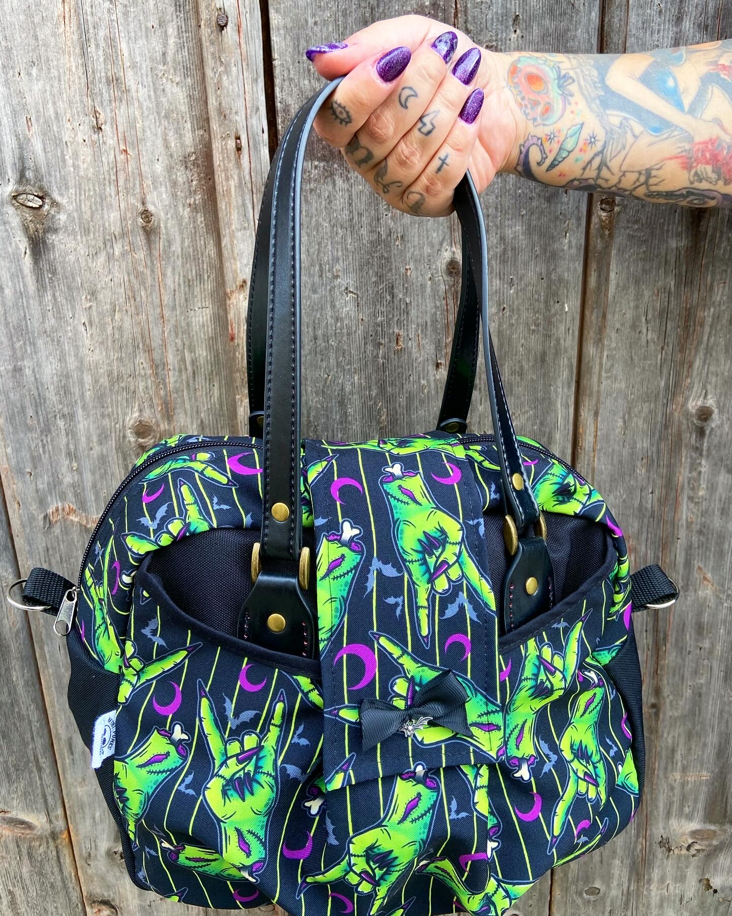 Mala de tecido preta com padrão de mãos verdes, luas lilás e alças pretas