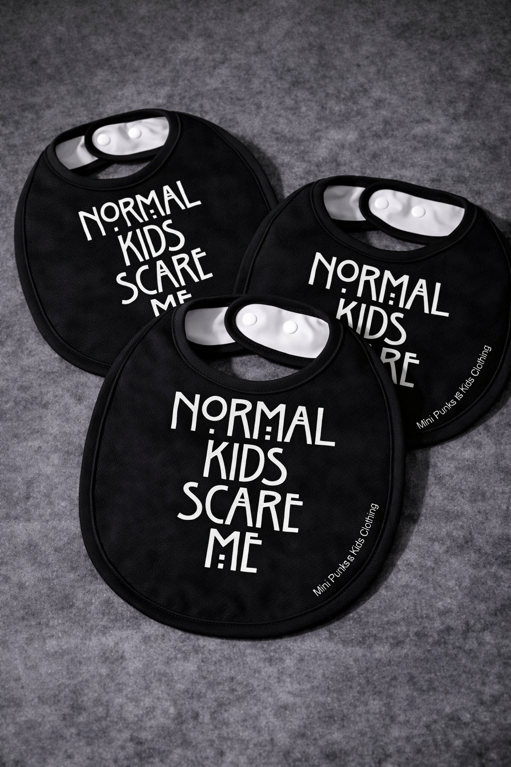 Três babetes pretos para bebés com texto branco e fecho de pressão.