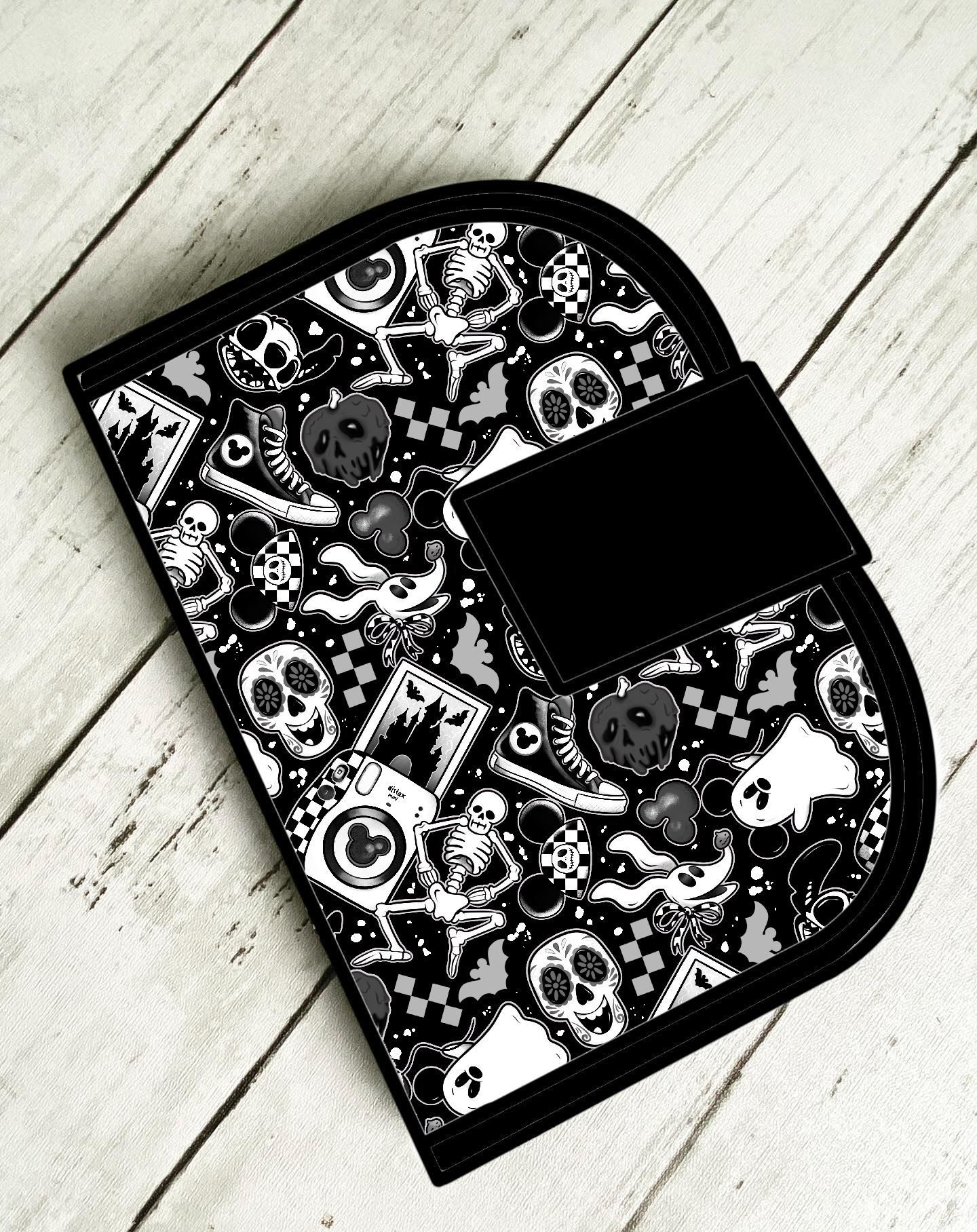 Bolsa preta com padrão de esqueletos e caveiras em preto e branco com aba preta sobre piso de madeira claro