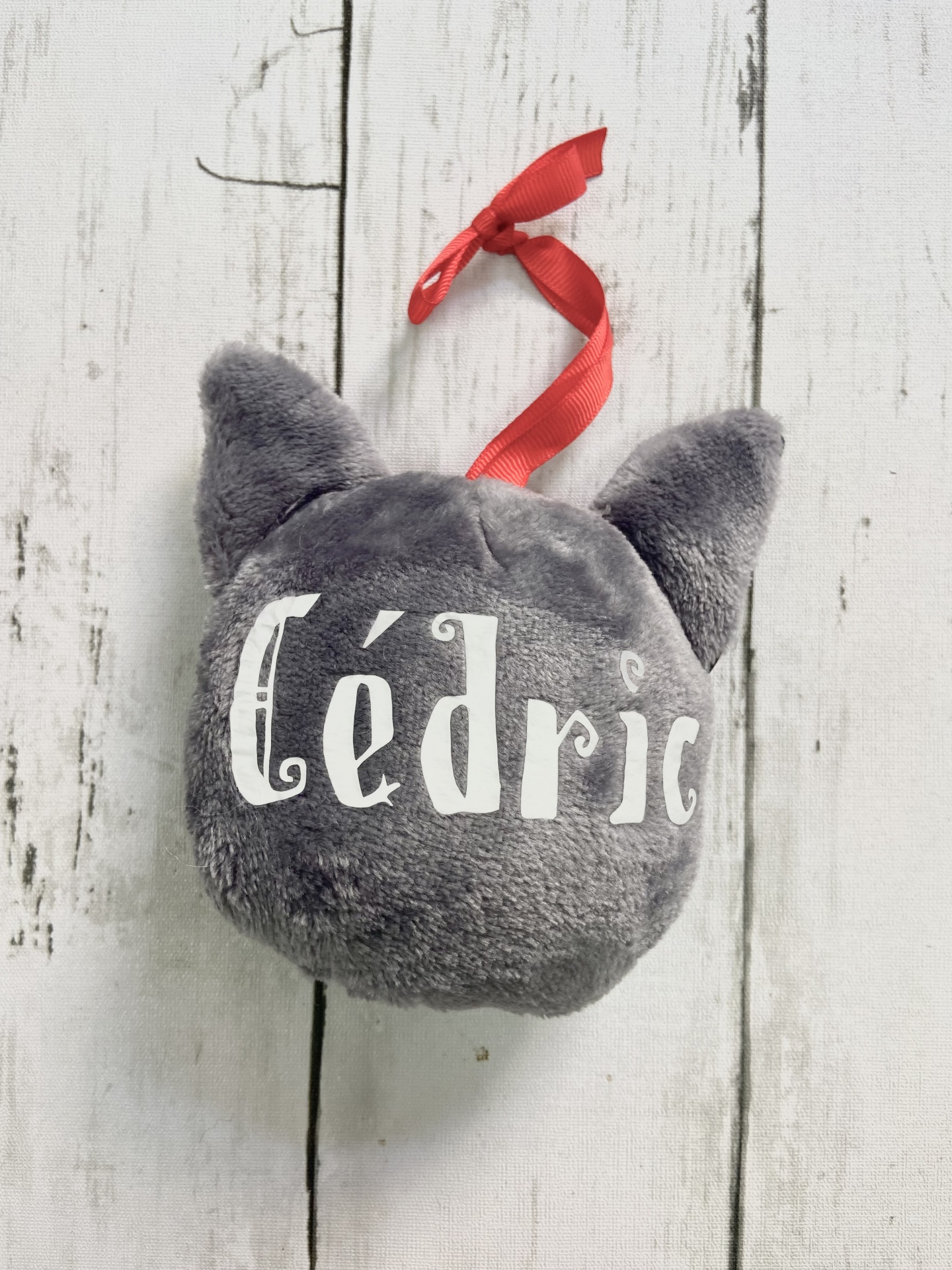 Pelúcia cinzenta em forma de cabeça de gato com nome 'cédric' e fita vermelha