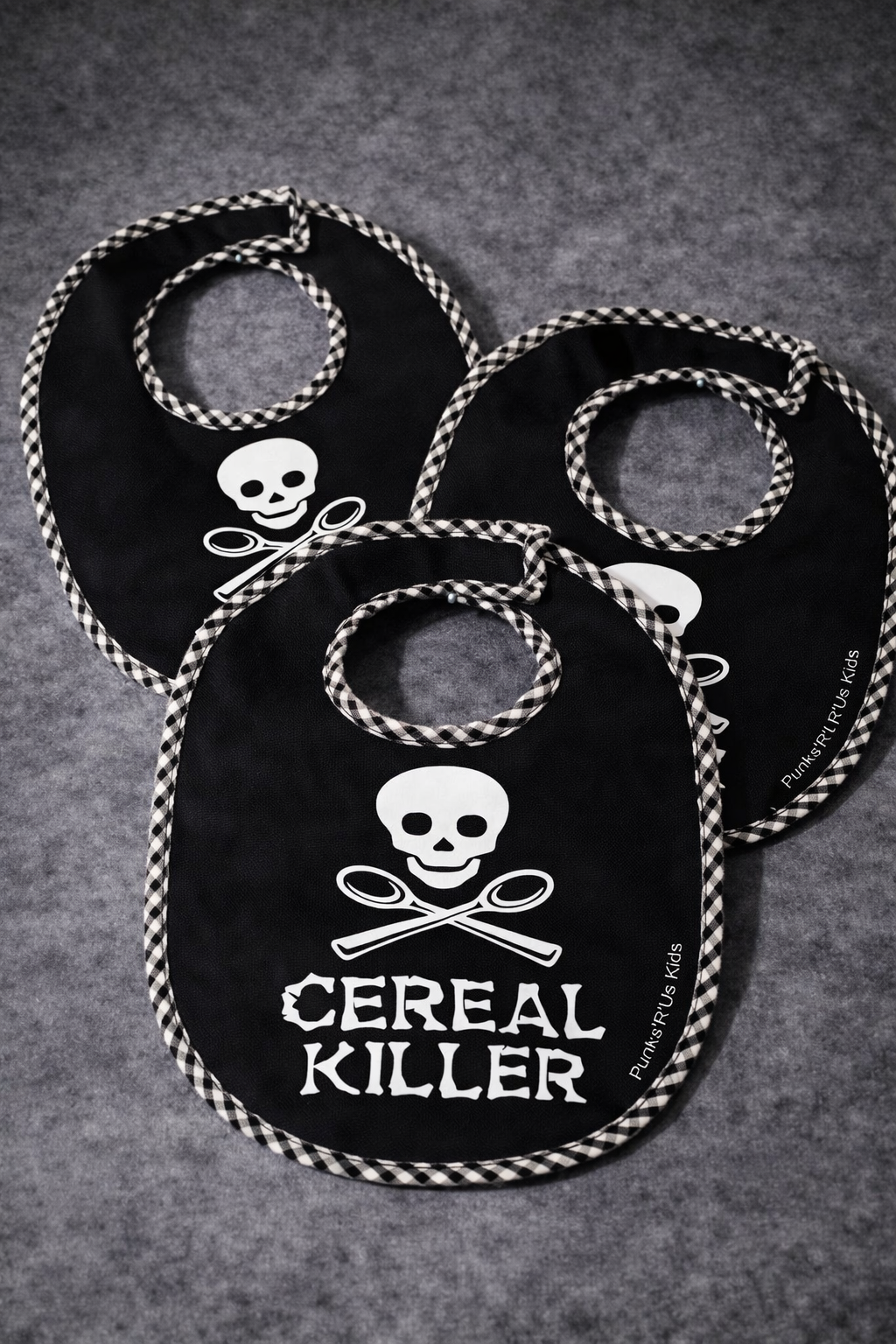 Três babetes pretos com padrão xadrez nas bordas e design de crânio e talheres cruzados com texto CEREAL KILLER