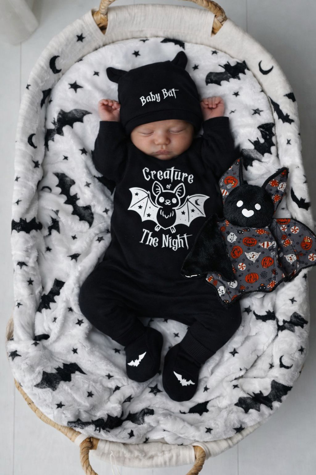 Bebé dormindo com roupa e gorro preto com texto, manta branca com morcegos e peluche morcego
