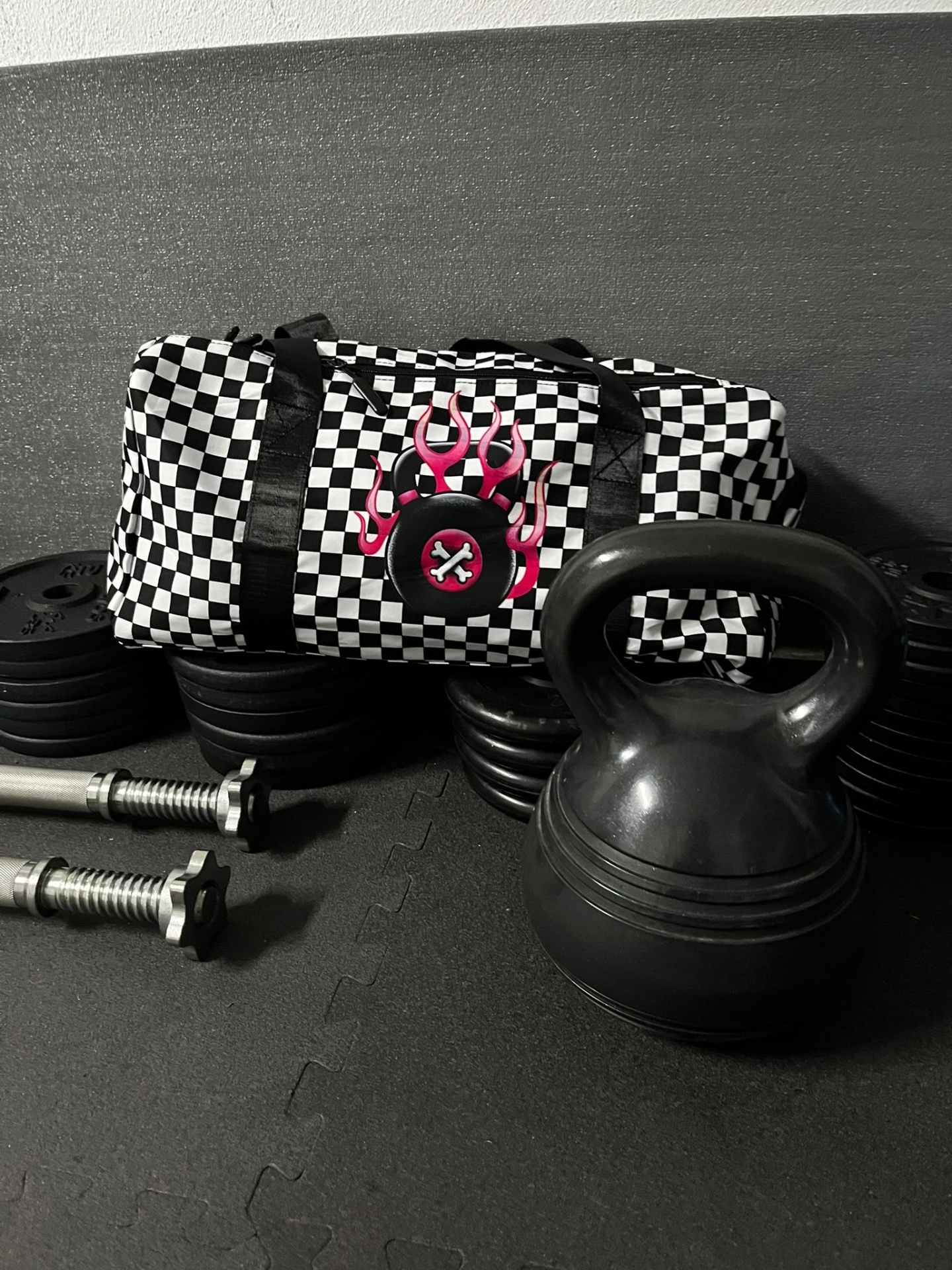 Saco de ginásio xadrez preto e branco com símbolo de fogo rosa, kettlebell preto e pesos de halteres em chão preto