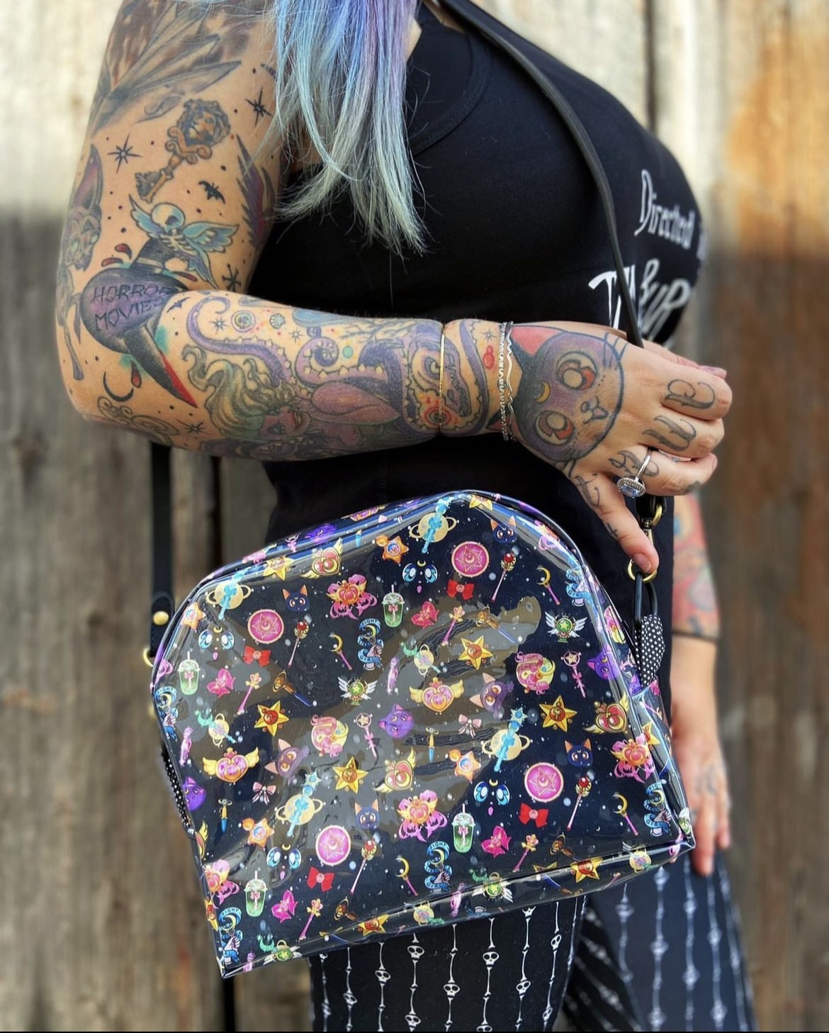 Bolsa quadrada em plástico translúcido com padrão colorido lúdico, segura por pessoa tatuada e vestida de preto