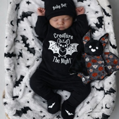 Bebé dormindo com roupa e gorro preto com texto, manta branca com morcegos e peluche morcego