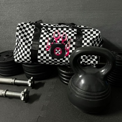 Saco de ginásio xadrez preto e branco com símbolo de fogo rosa, kettlebell preto e pesos de halteres em chão preto