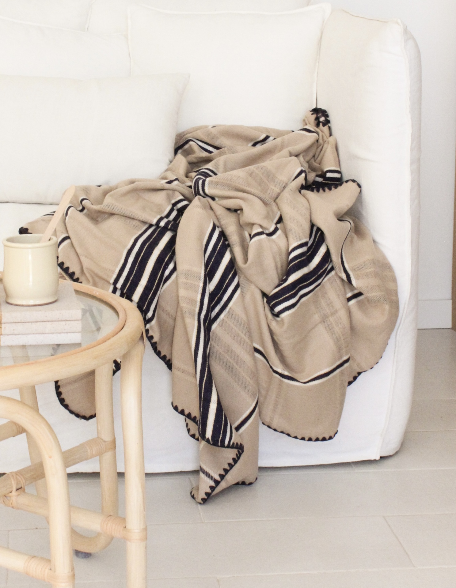 Manta bege com riscas pretas e brancas sobre sofá branco e mesa de madeira clara com caneca.
