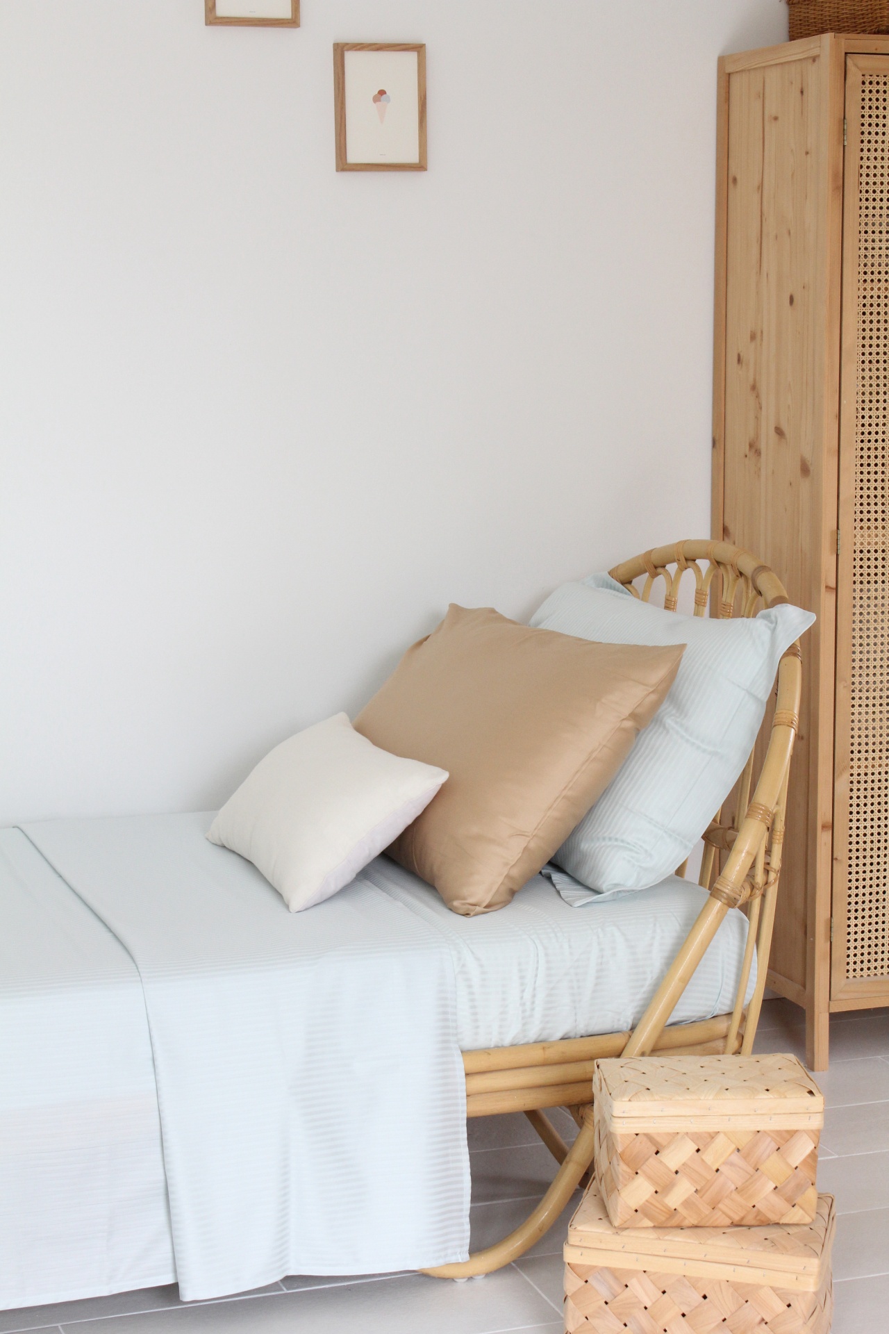 Cama de solteiro com roupa de cama azul clara e almofadas bege e branca junto a móvel de madeira e caixas de vime