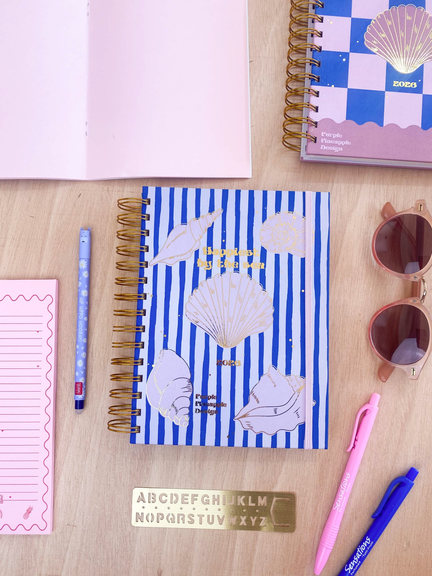 Caderno azul com riscas brancas e conchas douradas com materiais de escrita sobre mesa de madeira