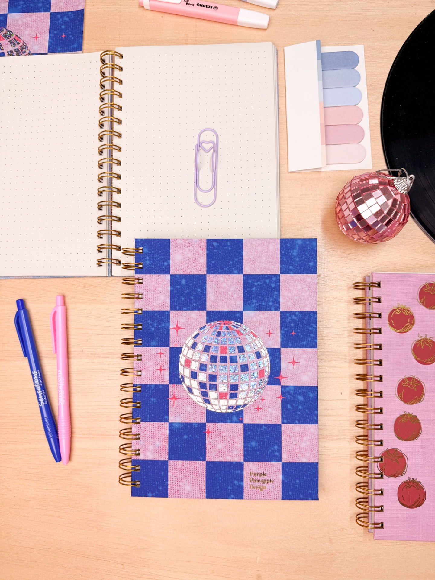 Cadernos espiral com capas decoradas em padrão xadrez e imagens de tomates, canetas, marcadores de página pastel e objeto decorativo rosa numa mesa de madeira.