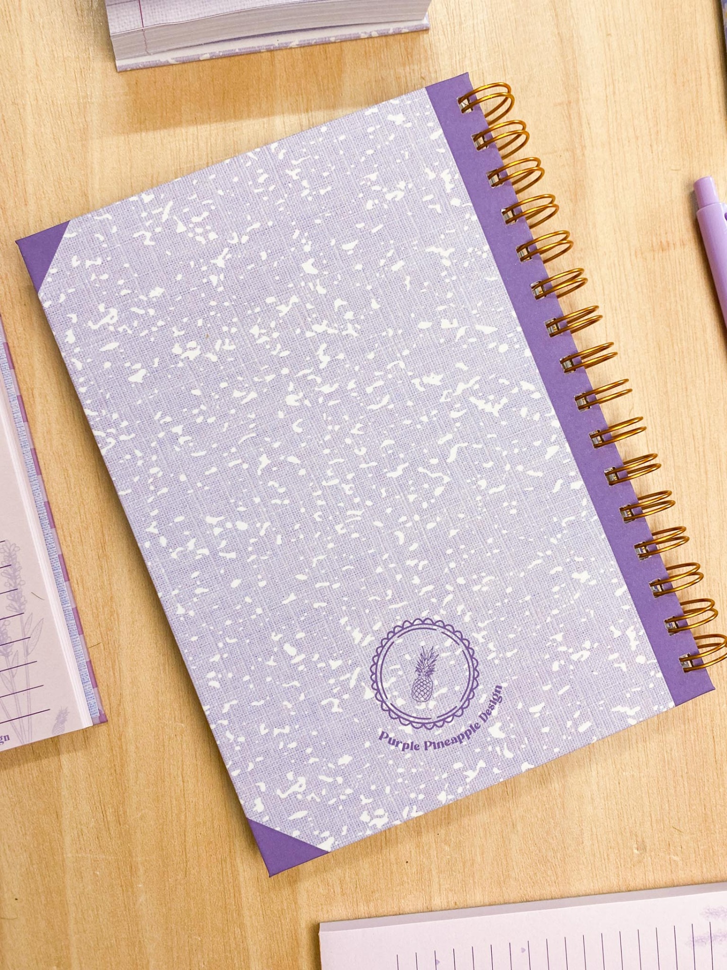 Caderno lilás com argolas e logótipo Purple Pineapple numa mesa de madeira