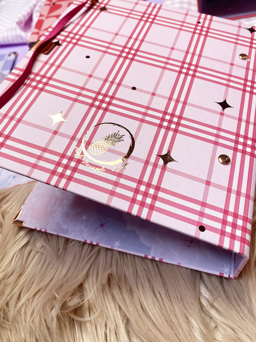 Caderno xadrez rosa e vermelho com abacaxi dourado sobre pelúcia bege