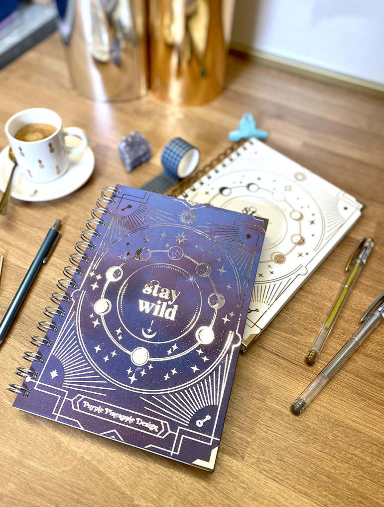 Dois cadernos espiralados com design celestial, canetas, café e fitas adesivas sobre mesa de madeira