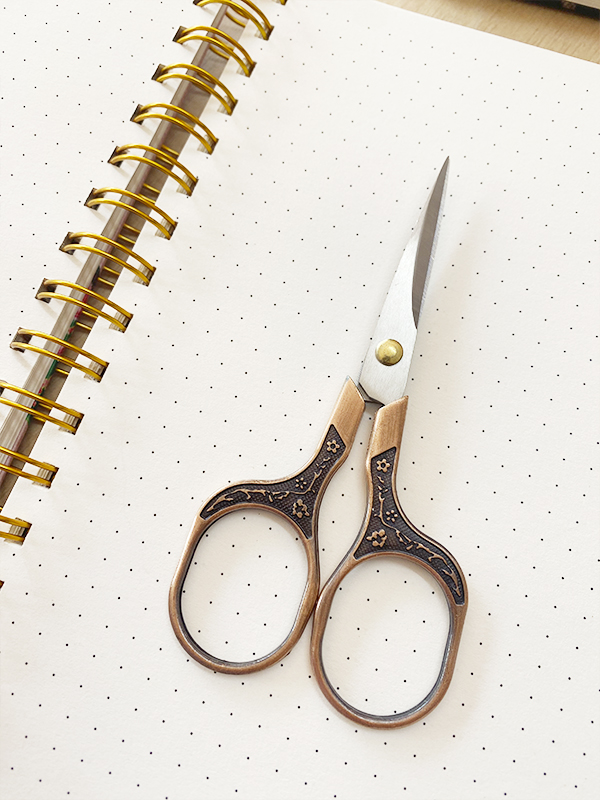 Tesoura pequena com pega em madeira decorada sobre página pontilhada de caderno.
