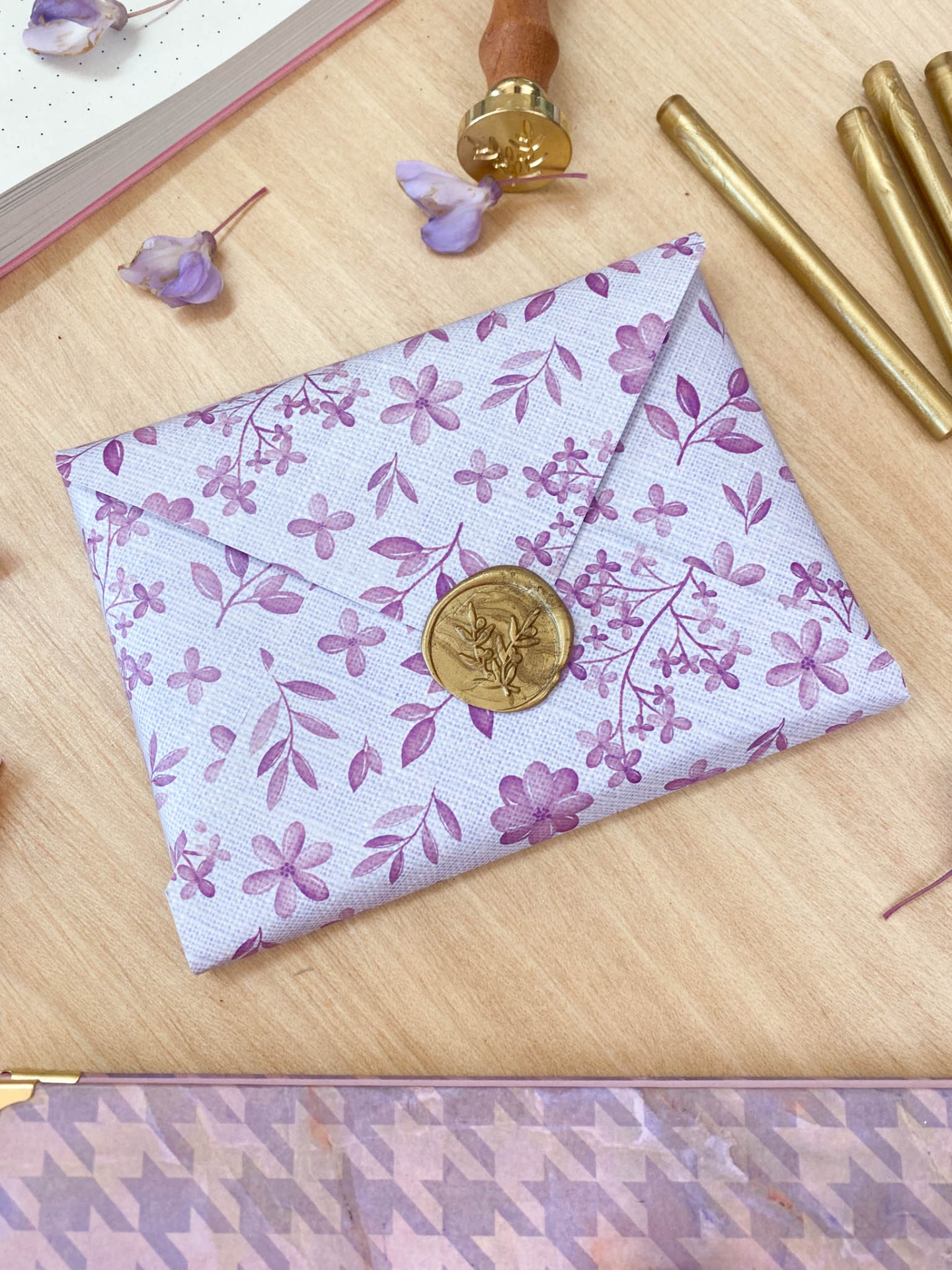 Envelope de tecido lilás com selo de cera dourado sobre mesa de madeira