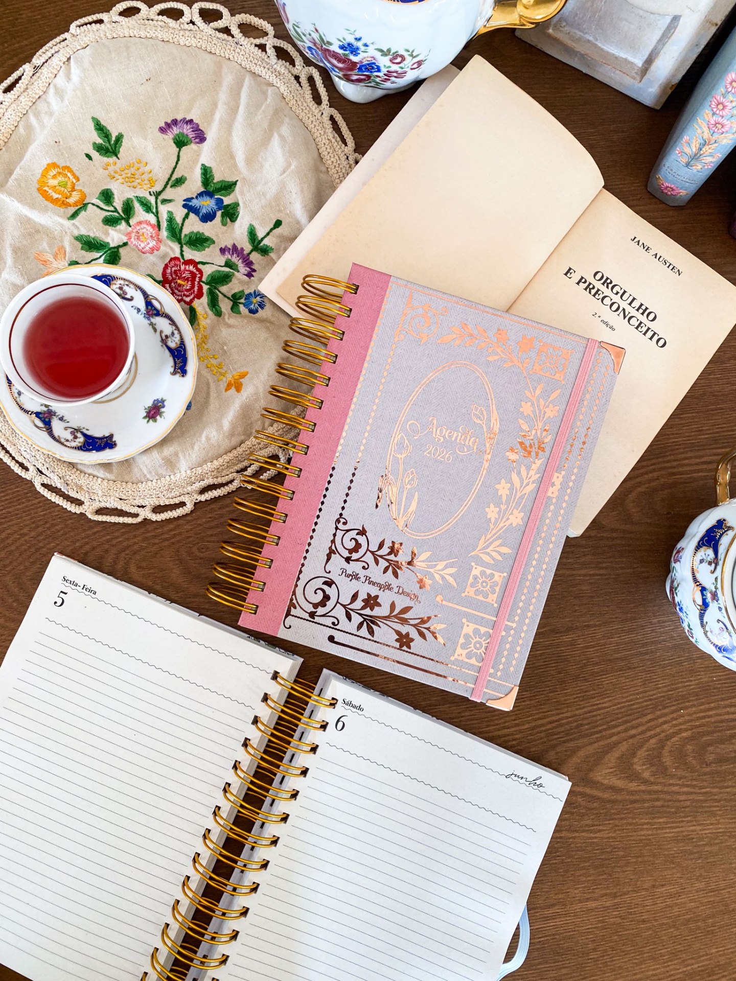 Agenda rosa e cinza com detalhes dourados ao lado de livro e chávena de chá em mesa de madeira