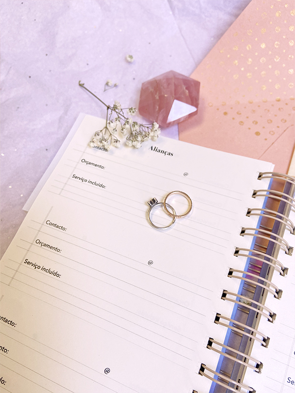 Dois anéis prateados estão sobre um caderno aberto com texto em português e elementos decorativos em fundo lilás e rosa.