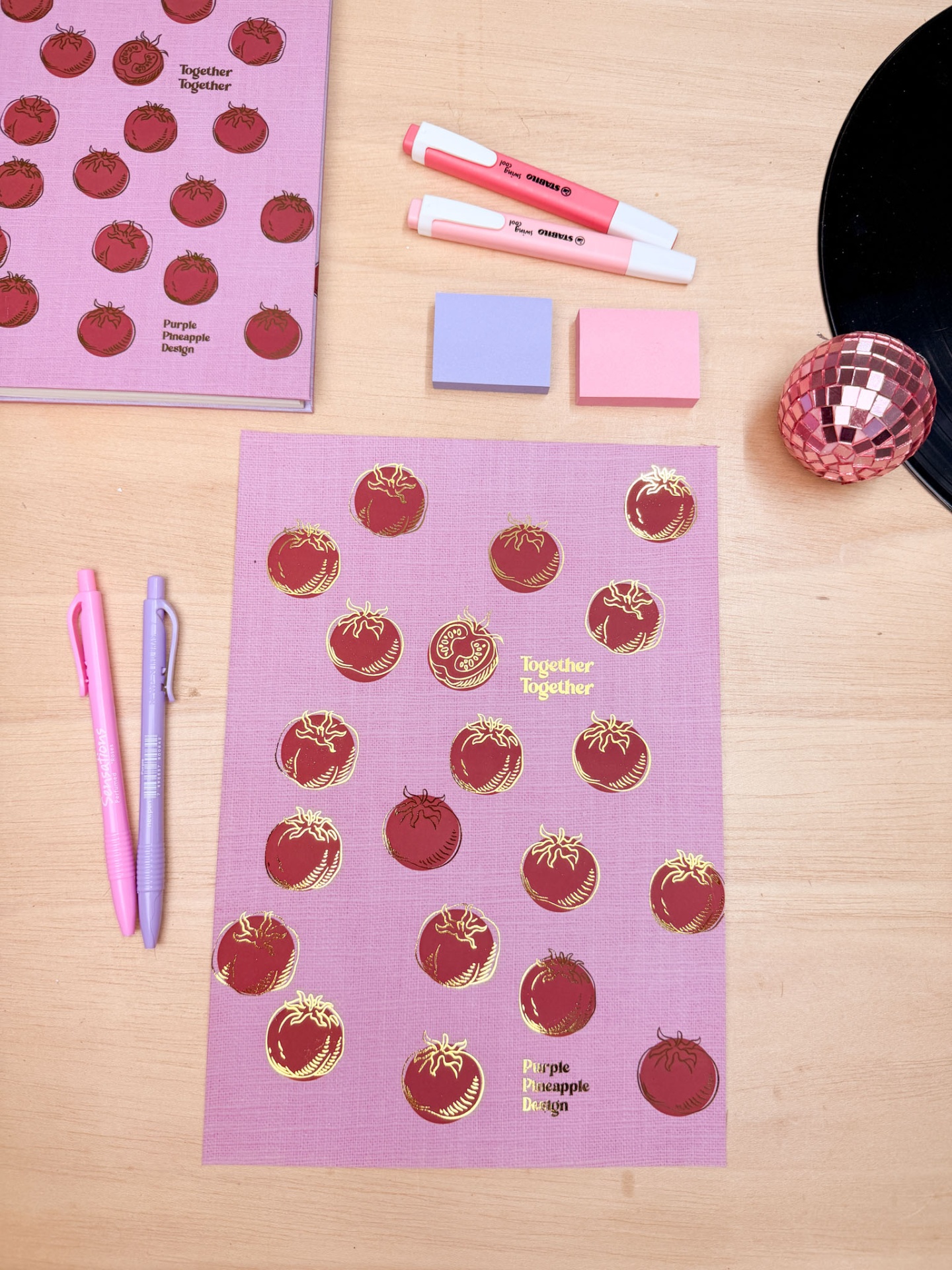 Conjunto de papelaria cor-de-rosa com desenhos de tomates e textos em dourado numa mesa de madeira